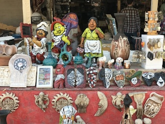 An eclectic collection of decorative items displayed on a storefront. The assortment includes colorful figurines, ornate masks, pottery, and various souvenirs. A clock and framed pictures can also be seen among the items, along with earrings and bracelets displayed on stands.