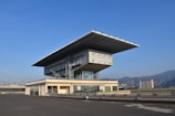 A modern building with a large overhanging roof structure and glass walls is situated on a rooftop with hills and a cityscape in the background. The clear sky enhances the architectural design, showcasing the blend of industrial elements and contemporary architecture.