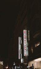 A nighttime scene featuring a tall building with illuminated signage displaying the words 'AVION' and 'HOTEL' in bright red letters vertically. The surrounding area is dimly lit, with other buildings partially visible in the background.