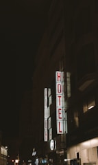 A nighttime scene featuring a tall building with illuminated signage displaying the words 'AVION' and 'HOTEL' in bright red letters vertically. The surrounding area is dimly lit, with other buildings partially visible in the background.