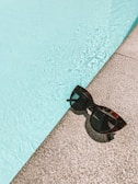 A pair of stylish sunglasses with tortoiseshell frames rests on a textured stone ledge beside a clear blue swimming pool, showcasing the contrast between the smooth lenses and the rippling water.