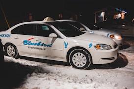 Two white taxis are parked on a snowy street at night. The vehicles have the name 'Bluebird Stephenville' written on the sides in blue lettering, with the numbers 9 and 1 visible. The scene is dimly lit, with a few lights visible in the background.