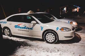 Two white taxis are parked on a snowy street at night. The vehicles have the name 'Bluebird Stephenville' written on the sides in blue lettering, with the numbers 9 and 1 visible. The scene is dimly lit, with a few lights visible in the background.