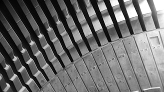 A black and white image showcasing the curved lines and geometric pattern of a large metal structure, possibly part of a turbine or industrial machinery. The image highlights the intricate design and texture of the metal, with various circular and rectangular indentations visible on the surface.