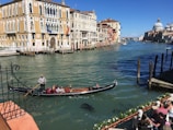 A gondola with a gondolier and passengers glides along a canal lined with historic, colorful buildings. People are sitting along the water's edge, enjoying the view under a clear blue sky. The architecture is ornate, with arched windows and intricate facades.