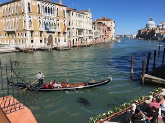 A gondola with a gondolier and passengers glides along a canal lined with historic, colorful buildings. People are sitting along the water's edge, enjoying the view under a clear blue sky. The architecture is ornate, with arched windows and intricate facades.