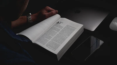 A focused adult reading a book with a look of determination, surrounded by notes and a laptop.