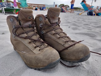 A pair of worn brown hiking boots is placed on a sandy beach, with people relaxing in the background. Various beach chairs and umbrellas are scattered around, with a wooden fence and some greenery visible further back. The boots are untied and slightly leaning against each other.