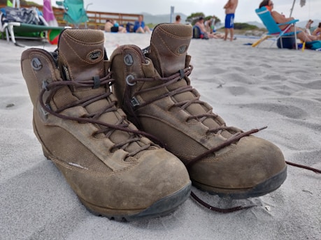 A pair of worn brown hiking boots is placed on a sandy beach, with people relaxing in the background. Various beach chairs and umbrellas are scattered around, with a wooden fence and some greenery visible further back. The boots are untied and slightly leaning against each other.