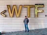 A person wearing sunglasses and a jacket stands in front of a large wall with the letters 'WTF' in gold. The phrase 'Where's The Food?' is seen above the letters. The wall has a neutral-toned, tiled appearance, and the floor is made of stone tiles.