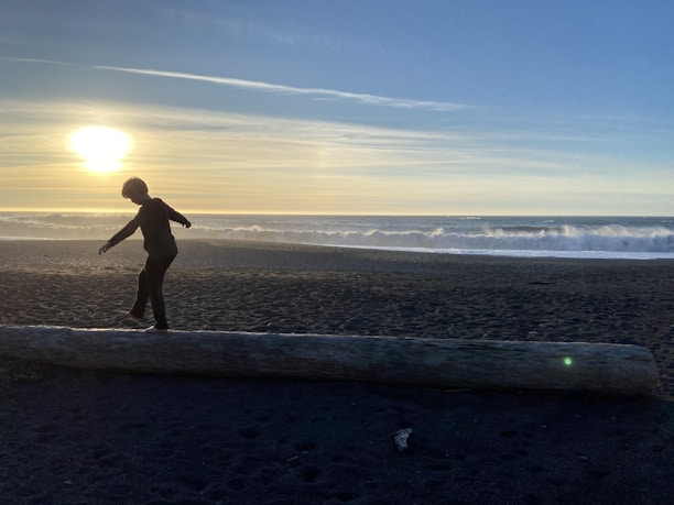 A person is balancing on a large log on a beach at sunset. The ocean waves are gently crashing in the background, and the sky is filled with soft hues of blue and orange as the sun nears the horizon.
