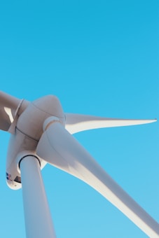 A close-up view of a large wind turbine against a clear blue sky. The image focuses on the turbine's blades and hub, capturing the sleek, aerodynamic design.