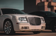 A luxury car with a distinctive grille and shiny rims is parked in an urban setting. The lighting suggests late afternoon or early evening, casting shadows that enhance the car's sleek design. The background includes part of a building, contributing to a sophisticated urban atmosphere.