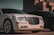 A luxury car with a distinctive grille and shiny rims is parked in an urban setting. The lighting suggests late afternoon or early evening, casting shadows that enhance the car's sleek design. The background includes part of a building, contributing to a sophisticated urban atmosphere.
