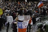 A large crowd gathers in a public square, participating in a protest. Many individuals hold signs with political and social messages, while some wave the Chilean flag. The atmosphere appears tense, with people engaged in animated discussions and chants. Several riot police officers in protective gear stand at the forefront, facing the protestors.