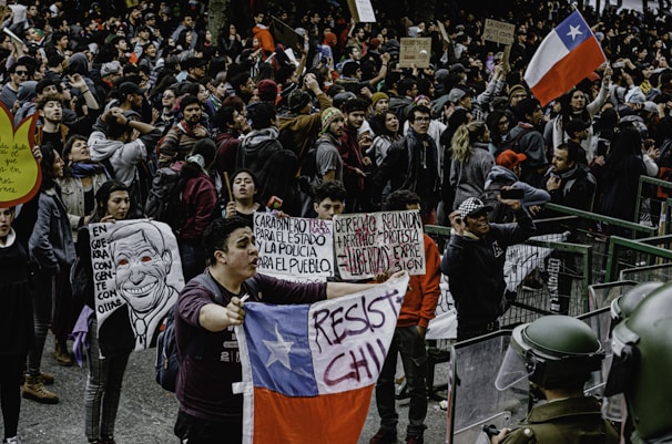 A large crowd gathers in a public square, participating in a protest. Many individuals hold signs with political and social messages, while some wave the Chilean flag. The atmosphere appears tense, with people engaged in animated discussions and chants. Several riot police officers in protective gear stand at the forefront, facing the protestors.
