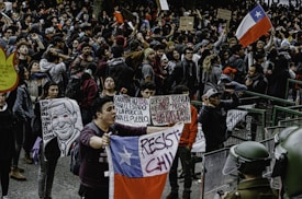 A large crowd gathers in a public square, participating in a protest. Many individuals hold signs with political and social messages, while some wave the Chilean flag. The atmosphere appears tense, with people engaged in animated discussions and chants. Several riot police officers in protective gear stand at the forefront, facing the protestors.