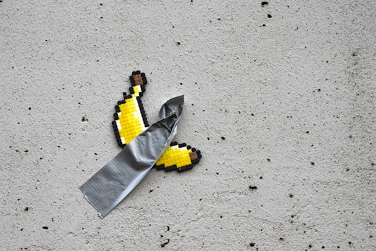 A pixelated banana peel is affixed to a concrete wall with a strip of duct tape. The banana appears to be made of small, cube-like pieces arranged to form a pixel art effect, while the tape is adhering it at an angle.