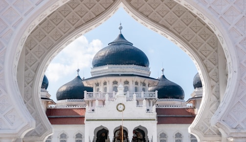 An ornate and intricately designed mosque with prominent black domes framed by a decorative white arch. The architecture features detailed patterns and a central clock above the entrance.