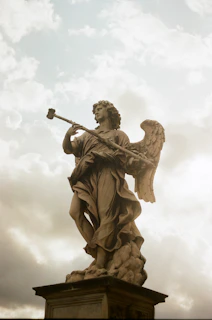 A marble statue of an angel standing on a pedestal, captured against a backdrop of a cloudy sky with soft lighting. The angel is depicted with large wings and holding an object resembling a spear or staff.
