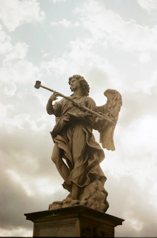 A marble statue of an angel standing on a pedestal, captured against a backdrop of a cloudy sky with soft lighting. The angel is depicted with large wings and holding an object resembling a spear or staff.