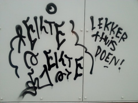 Graffiti text is displayed on a light-colored background. The writing is in a stylized, bold, black font with some elements appearing abstract or decorative.