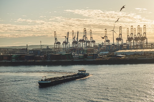 An industrial harbor scene with multiple cranes lining the waterfront and a large barge moving along the water. Seagulls are flying in the sky above, and a wind turbine can be seen in the distance. The setting sun casts a warm glow over the scene, highlighting the calm water and the activities of the port.
