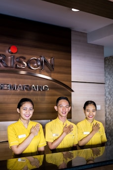 Three individuals are standing behind a reception desk, each wearing a yellow uniform. They are smiling and have their right hands placed over their chests. The background features a wooden wall with part of a logo and the word 'SEMARNG' visible.