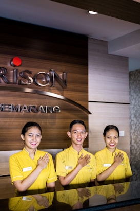 Three individuals are standing behind a reception desk, each wearing a yellow uniform. They are smiling and have their right hands placed over their chests. The background features a wooden wall with part of a logo and the word 'SEMARNG' visible.