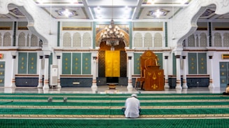 An ornate mosque interior with intricate designs featuring arches and decorative panels. The room has multiple prayer carpets laid out on the floor, and a person is seated in the middle of the carpeted area. A large wooden pulpit, chandeliers, and a golden ornamental area are visible.
