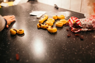 Several pieces of yellow dough, some shaped into swirls and others with raisins, are arranged on a black countertop. A hand is reaching towards the dough, and an open bag of raisins sits nearby with some scattered raisins.