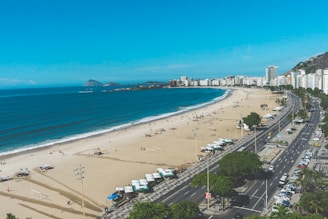An expansive beach stretches alongside a bustling street, with the ocean forming gentle waves on the sand. High-rise buildings line the shore in the distance, while numerous small tents and umbrellas dot the beach. People are scattered across the sand, engaging in various beach activities, and vehicles are parked along the decorated sidewalk.