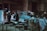 An industrial workshop setting with two people interacting near a vintage car chassis. The space is cluttered with various tools, equipment, and makeshift tables. Shopping carts filled with miscellaneous items, such as a jerry can and cloth, are scattered around. The environment is dimly lit, with a predominantly teal and grey color scheme.