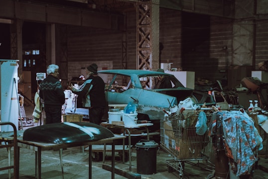 An industrial workshop setting with two people interacting near a vintage car chassis. The space is cluttered with various tools, equipment, and makeshift tables. Shopping carts filled with miscellaneous items, such as a jerry can and cloth, are scattered around. The environment is dimly lit, with a predominantly teal and grey color scheme.