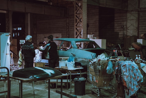 An industrial workshop setting with two people interacting near a vintage car chassis. The space is cluttered with various tools, equipment, and makeshift tables. Shopping carts filled with miscellaneous items, such as a jerry can and cloth, are scattered around. The environment is dimly lit, with a predominantly teal and grey color scheme.