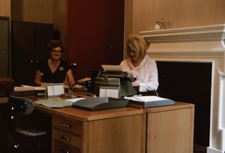 Two women are seated in an office environment, working at a large wooden desk. One woman on the right is typing on a typewriter, while the other woman on the left is sitting with papers in front of her. The office has a traditional aesthetic with a neutral color palette, including a dark filing cabinet in the background and decorative molding on the walls.