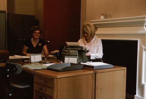 Two women are seated in an office environment, working at a large wooden desk. One woman on the right is typing on a typewriter, while the other woman on the left is sitting with papers in front of her. The office has a traditional aesthetic with a neutral color palette, including a dark filing cabinet in the background and decorative molding on the walls.