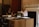 Two women are seated in an office environment, working at a large wooden desk. One woman on the right is typing on a typewriter, while the other woman on the left is sitting with papers in front of her. The office has a traditional aesthetic with a neutral color palette, including a dark filing cabinet in the background and decorative molding on the walls.