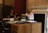 Two women are seated in an office environment, working at a large wooden desk. One woman on the right is typing on a typewriter, while the other woman on the left is sitting with papers in front of her. The office has a traditional aesthetic with a neutral color palette, including a dark filing cabinet in the background and decorative molding on the walls.