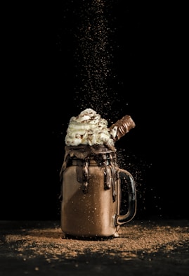 A decadent milkshake in a mason jar is overflowing with whipped cream and drizzled with chocolate. A chocolate bar is inserted into the top, and cocoa powder is artistically sprinkled, capturing a dynamic moment against a dark background.