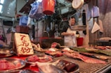 A market stall with various cuts of meat displayed on metal trays and a table. The vendor, wearing a white apron, stands behind the counter. A weighing scale is suspended above the counter. The setting is indoors with numerous plastic bags and containers hanging and stored in the background.