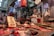 A market stall with various cuts of meat displayed on metal trays and a table. The vendor, wearing a white apron, stands behind the counter. A weighing scale is suspended above the counter. The setting is indoors with numerous plastic bags and containers hanging and stored in the background.