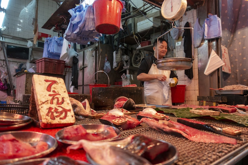 A market stall with various cuts of meat displayed on metal trays and a table. The vendor, wearing a white apron, stands behind the counter. A weighing scale is suspended above the counter. The setting is indoors with numerous plastic bags and containers hanging and stored in the background.