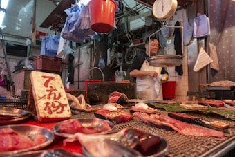 A market stall with various cuts of meat displayed on metal trays and a table. The vendor, wearing a white apron, stands behind the counter. A weighing scale is suspended above the counter. The setting is indoors with numerous plastic bags and containers hanging and stored in the background.
