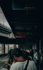 A group of people waits under a covered platform beside railway tracks. The platform is sheltered by a metal roof, and there is a digital clock displaying the time 6:24:42. Some people are standing while others appear to be looking at their phones, giving the scene a sense of movement and anticipation.