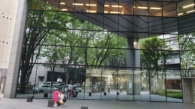 A large glass building reflects the surrounding urban environment, including trees and people walking by. The windows show a clear view of the interior lights and structural design of the building. Outside, a few people are gathered, some sitting and some walking. The reflection creates a visually interesting scene, merging the natural and built environments.