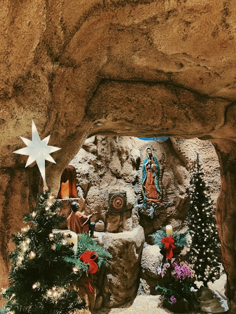 A grotto-like setting featuring a religious statue mounted on a rocky wall. There are decorated Christmas trees with lights and red bows on either side. A star-shaped light hangs on the left. The atmosphere is serene and reverent, with additional decorative elements including a candle and small potted plants.
