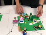 Hands are assembling a small Lego model on a green baseplate. The model includes various colorful Lego pieces such as minifigures, flowers, grass, a cake-like structure, and a vehicle.