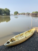 A serene riverside scene with a yellow kayak resting on a pebbled shore next to calm water. In the background, lush green trees line the bank and a few small piers and buildings are visible across the river. The sky is clear and blue, adding to the tranquil atmosphere.