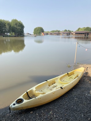 A serene riverside scene with a yellow kayak resting on a pebbled shore next to calm water. In the background, lush green trees line the bank and a few small piers and buildings are visible across the river. The sky is clear and blue, adding to the tranquil atmosphere.